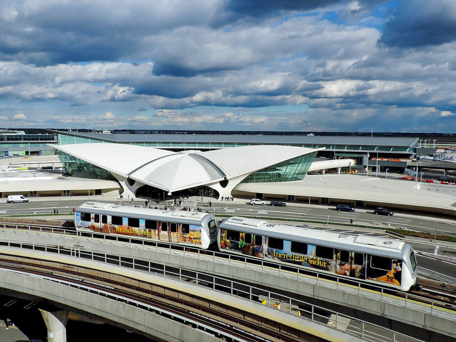 ขบวนรถไฟ AirTrain JFK สีขาวคาดแถบสีน้ำเงินและเหลือง กำลังวิ่งอยู่บนรางยกระดับ โดยมีฉากหลังเป็นท้องฟ้าและป้ายบอกทางบนทางด่วน